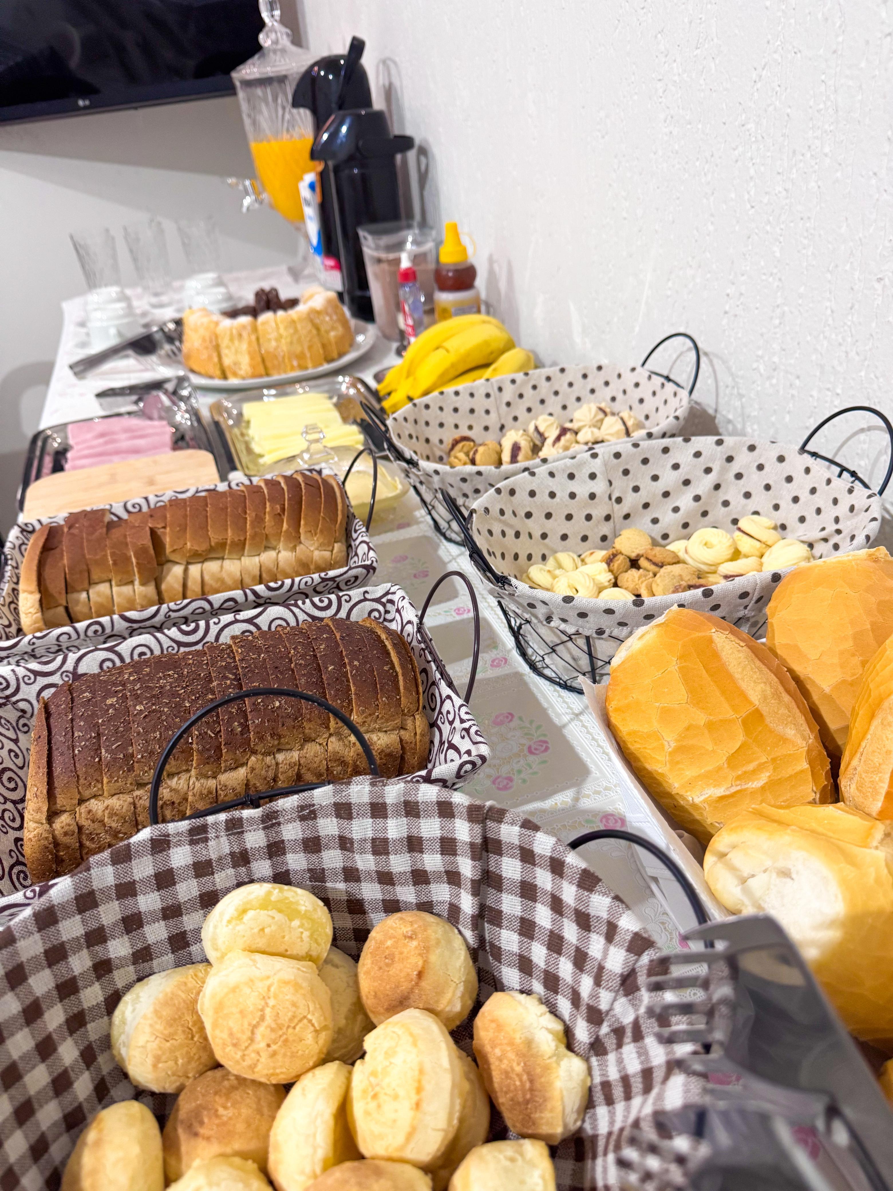 Mesa de café da manhã vista de lado, com pães, bolo e garrafas térmicas